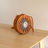 A small, woven crossbody bag made from rattan features real seashell embellishments and a star-like pattern on its front. The brown-colored bag has an adjustable strap for comfortable wear and is adorned with visible seashells. It's placed on a wooden surface in a casual environment, evoking coastal or summer themes.