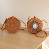 Two woven bags, one larger than the other, rest on a light-colored surface against a plain wall. The front of each bag features a circular design with a star-like pattern within. Brown handles adorned with seashells connect to both bags. The image showcases the intricate details and natural materials used in their construction.