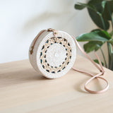 A woven crossbody bag with a floral pattern rests on a light-colored surface, made of rattan with a circular design and adjustable pink strap. The bag features delicate white floral details and a compact size, exuding timeless elegance.