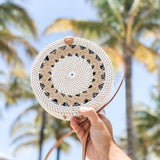 A person holds up a round woven straw crossbody bag, showcasing its beige color and white and pink floral patterns against a tropical backdrop of palm trees and a clear sky. The bag features a circular shape with a handle on top, made from natural materials with a casual yet stylish design.