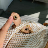 A person's hand holds a wicker crossbody bag, showcasing its circular shape with a flap closure. The bag is made of a light-colored woven material, possibly rattan or bamboo, featuring two brown leather straps and a metal button. The background suggests an outdoor setting, likely in a store or at home.