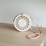 A woven crossbody bag, made from rattan with a floral white pattern, rests on a surface. The bag features a circular shape, adjustable pink leather strap, and intricate weaving details.