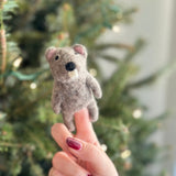 A light brown needle felted beaver finger puppet, crafted from 100% wool, is held by a female hand with dark red nail polish against a softly blurred Christmas tree background. This handmade felt beaver finger puppet features a black nose and small ears, showcasing its delightful 3D design.