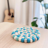 A round sky-blue felt ball pot trivet, handcrafted from ethically sourced 100% organic lamb wool, rests on a light wooden surface. This unique handmade coaster, featuring alternating blue, white, and grey felt balls, is carefully arranged in a circular pattern. The elegant and cozy home decor piece is ethically made by Nepalese female artisans, with a blurred guitar and potted plant in the background.