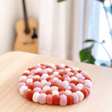 A round felt ball pot trivet rests on a light wood-grain surface, bathed in natural light. This handcrafted organic lamb wool felt trivet features tightly packed, hand-stitched felt balls in shades of white, light pink, and coral, giving it a textured and dimensional appearance. Its artisanal quality adds a cozy touch, perfect for home decor.