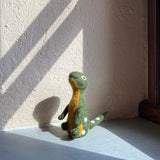 A handcrafted green wool felt Brontosaurus finger puppet sits in a sunlit corner, resting against a textured light beige wall with dark brown trim. This playful felted Brontosaurus puppet features a yellowish-tan neck and underbelly, with small white spots along its back and tail. Its needle felted design includes a long neck, small arms, and tiny round eyes, inviting imaginative play for kids.