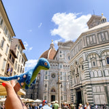 A vibrant, handcrafted wool felt Brontosaurus finger puppet, part of the Dino Brothers collection, is held by a female hand against a sunny European cityscape. This unique needle-felted Brontosaurus puppet features a long neck and white spots on its back, with a lifelike 3D design. Its vivid blue and green colors create a whimsical contrast, evoking joy and imagination.