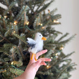 A handcrafted wool felt eagle finger puppet sits on a person's fingers, displayed against the soft, warm glow of a Christmas tree. This unique needle felted eagle puppet is predominantly white with a textured appearance, accented by a bright orange beak and feet, and distinctive black detailing on its wings.