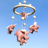 The wool felt baby mobile, featuring whimsical flying pigs, is suspended against a clear blue sky. It has a thick, circular off-white felt frame adorned with matching felt balls at varying heights. Four handcrafted pink wool felt pigs, detailed with white wings and facial stitching, hang below, appearing to float peacefully. This dreamlike scene is perfect for a baby's nursery.