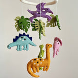 A whimsical Jurassic dinosaur felt baby mobile is presented against a plain, light-colored background. Felt dinosaurs and green monstera leaves are suspended by white strings from a white, felt-covered ring. It features a purple Pterosaur, blue Stegosaurus, green T-Rex, orange Brontosaurus, and pink Triceratops, each with unique markings, creating a playful scene.