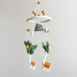 A wool felt llama baby mobile is displayed against a plain, light gray background. It features a white felt hoop adorned with tan balls. Hanging below are soft, handcrafted felt shapes including gray mountains, white leaves, green cacti with yellow flowers, and white llamas with orange blankets. This charming mobile creates a calming and gentle nursery ambiance.