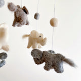 A felt baby mobile, featuring a central elephant figure made from wool felt, hangs from a string against a white background. The elephant appears to be swinging gently, surrounded by smaller felt animals that are not clearly distinguishable due to their size and distance from the camera. The overall mood is playful and whimsical, with emphasis on the soft, textured appearance of the felt animals.