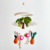 A colorful felt baby mobile, suspended from a white string, features a pineapple, two flamingos, and a palm tree crafted from wool felt. The tropical theme is playful and vibrant, with each element giving off a soft texture. A neutral background allows the colorful elements to stand out.