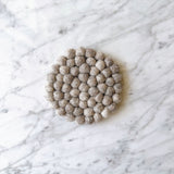 A single, round gray felt ball coaster is displayed on a white marble surface with subtle gray veining. This handcrafted felt coaster is composed of numerous tightly packed, hand-stitched felt gray balls made from 100% organic lamb wool. Its minimalist design, ethically handmade by Nepalese female artisans, evokes calm and understated elegance.