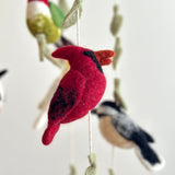A close-up of the wool felt bird baby mobile features a vibrant red wool felt cardinal hanging from a wooden frame. Other blurred felt birds like a hummingbird and chickadee, along with small sage-green felt leaves, are suspended by off-white strings against a soft white background, creating a cozy, playful atmosphere.