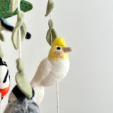 A charming felt bird baby mobile is shown in a close-up against a plain white background, highlighting its intricate details. Several handcrafted felt birds and decorative leaves hang from a natural wooden frame by white strings. One prominent bird features a yellow head and white body, while the felt leaves display visible stitching.