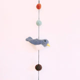 A colorful felt bird garland hangs vertically against a plain white background. It showcases a detailed 3D needle-felted Nuthatch bird with a white belly, yellow beak, and small black eye. Above the bird are light green and orange wool felt balls, with a brown felt ball below. The charming wool felt garland is suspended by a black string, ending with a small copper bell.