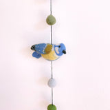 A charming wool felt bird garland hangs on a plain white wall. A close-up shows a meticulously crafted 3D blue tit bird with a blue head, yellow body, and black beak. Muted green and gray wool felt balls are strung above and below the bird on a black string, creating a whimsical display.