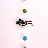A charming wool felt bird garland, featuring a detailed 3D needle felted woodpecker, hangs against a bright white wall. The woodpecker, predominantly white with black markings, boasts a red crest and small grey beak. It connects via a thin black string to green and teal felt balls, with a copper bell visible at the end of the garland, adding a playful touch.