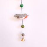 A handcrafted wool felt bird garland is displayed against a simple white backdrop. This charming felt garland features multiple birds, including a gray, white, and red Robin. It also includes green and gray wool felt balls strung along a black cord, finished with a copper bell at the bottom, adding a whimsical touch.