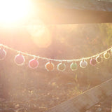 A colorful wool felt lightbulb garland is suspended outdoors in warm sunlight against blurred foliage and a rustic wooden structure. The garland features rounded, lightbulb-shaped felt balls in pink, yellow, green, and white, evenly spaced on a thin, natural fiber string. The warm sunlight gives the scene a dreamy, whimsical feel.