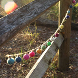 A whimsical wool felt garland, adorned with colorful felt balls resembling miniature lightbulbs, is strung across a weathered wooden fence in a bright outdoor setting. This approximately 4-foot garland features spherical pink, blue, green, yellow, and white felt balls, each with a small stem detail, connected by rustic twine.