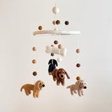 A Felt Dachshund dog baby mobile hangs against a neutral, off-white backdrop. It features four soft felt dachshunds in cream, khaki, brown, and black, suspended from a white felt frame. A white felt bone is centered above the dogs, creating a comforting aesthetic.