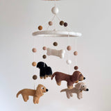 A felt Dachshund baby mobile, featuring four delightful dachshund dogs in cream, khaki, brown, and black, is displayed against a plain white background. The dogs and a white felt bone hang from a white felt frame, creating a calm, playful atmosphere.