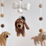 A charming felt Dachshund dog baby mobile is presented in a close-up against a simple off-white background. It features a brown felt Dachshund prominently, along with cream and khaki felt Dachshunds and a white felt bone. All are suspended by thin white strings from a white felt frame, creating a gentle and playful nursery display.