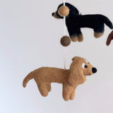 A felt Dachshund Dog Baby Mobile is displayed against a solid white background. It features four adorable felt Dachshunds in cream, khaki, brown, and black, all in profile with elongated bodies and stubby legs. A felt bone sits at the center, all suspended from a white felt frame. The figures have a visible, fuzzy texture, conveying a handmade, comforting feel ideal for a nursery.