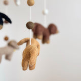 A khaki wool felt dachshund dog, part of a charming baby mobile, hangs suspended by a white string against a plain white background. Its rear faces the viewer, showcasing its detailed tail and hind legs. Blurred brown, cream, and black felt dachshunds, a white felt frame, and a central felt bone are visible behind it, creating a warm, playful nursery scene.