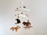 A felt Dachshund baby mobile is shown against a muted off-white background. Four felt dachshunds in cream, khaki, brown, and black hang from a white felt frame, arranged in a circular pattern. A white felt bone hangs playfully in the center.