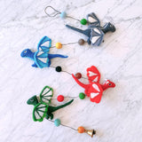 A charming wool felt dragon garland featuring four 3D felted pterosaurs is displayed on a white marble surface. The red, blue, green, and gray pterosaurs have white accents on their wings and heads and are suspended from a black string. Interspersed wool felt balls in shades of brown, green, yellow, and blue, and a copper bell at the end, complete this approximately 4-foot long nursery garland.