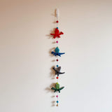 A colorful felt dragon garland is displayed against a plain off-white wall. The garland features four 3D felt pterosaurs in red, blue, gray, and green. They are strung along with colorful felt balls, and a small copper bell hangs at the bottom.