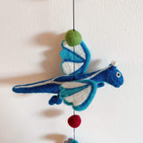 A vibrant blue felt dragon from a felt dragon garland is suspended by a dark string against a clean white wall. Crafted from pure wool felt, the playful dragon features shades of blue and white on its wings and body, adding a cheerful touch.