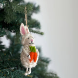 A cream-colored Felt Easter Bunny Sheep Ornament made of wool felt hangs on a Christmas tree. This unique felted ornament features a fluffy sheep with long bunny ears that have pink insides, a stitched pink nose, and a black stitched smile. The sheep ornament charmingly holds two orange felted carrots with green tops, with a jute string visible for hanging.