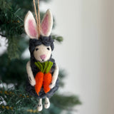 The wool felt Easter bunny sheep ornament hangs from a jute string on a tree, against a softly blurred evergreen background. This playful needle felted sheep features a grey wool body with white feet, a light pink nose, and black eyes. It is adorned with white bunny ears with pink insides and holds two orange carrots with green tops.