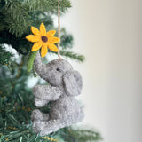 A gray wool felt elephant ornament, with a textured body and floppy ears, hangs from the green branches of a Christmas tree. The adorable elephant holds a yellow flower with a brown center and green stem in its trunk. A jute string allows this charming felt Christmas ornament to be suspended, creating a cozy and festive atmosphere.