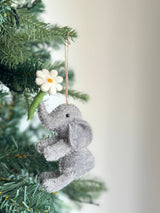 A charming handmade wool felt elephant ornament, adorned with a flower, hangs gracefully on a lush green Christmas tree. This gray felt elephant features a cheerful white flower with a yellow center held in its trunk. It has stitched details for its eyes and a natural jute string for easy hanging, blending seamlessly with the festive setting.