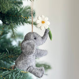 A handcrafted felt elephant with flower ornament hangs gracefully from a green Christmas tree branch. This soft gray felt elephant ornament features a black eye, meticulously shaped ears, and a trunk playfully holding a white felt flower with a yellow center and green stem. A jute string allows it to hang beautifully.