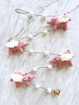 A whimsical wool felt flying pig garland is displayed on a white marble surface. It features four pink wool felt pig figures, each adorned with white wool felt wings, strung along a black string. White wool felt balls are interspersed between the pigs, completing the playful design.