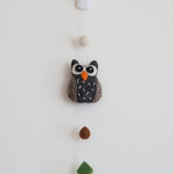 A wool felt forest animal garland is displayed against a plain white wall, suspended from a subtle white hook. The handmade garland features a detailed 3D owl with a gray body, brown accents, white stitching, and an orange beak. Above the owl is a white felt ball, while a green felt tree hangs below, connected by a thin string. A small copper bell completes the natural, handcrafted design.