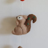 A light brown, 3D needle-felted squirrel from a felt forest animal garland hangs against a plain white wall. This 100% wool squirrel features a darker brown tail adorned with small green and red embroidered details. It is suspended by a thin white string, with a red felt element above and a small brown felt ball below, suggesting the garland extends to a copper bell.