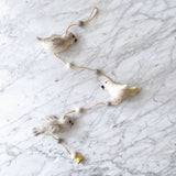 A felt seal garland is displayed on a white marble surface, exuding a calm and playful atmosphere. The garland features three soft felt seals—two light grey and one white—interspersed with alternating white and grey felt balls on a brown string. The bottom felt seal is adorned with a small copper bell.