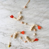 A playful Felt Shrimp Garland rests on a clean white marble surface. It features three white felt shrimp with light tan stripes and bright orange tails, evenly spaced along a thin twine. Small, round felt balls in light pink and white are interspersed between the shrimp, and a single copper bell adorns one end.