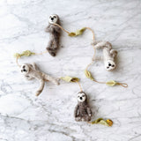 A whimsical felt garland, featuring four grey and brown sloths and light green felt leaves, rests on a white marble surface. This serene sloth felt garland showcases sloths in various playful poses, including hanging upside down, meditating, reaching out, and standing upright, all connected by thin twine.