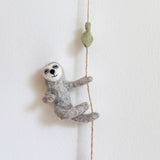 A whimsical sloth felt garland hangs against a stark white backdrop. A handcrafted grey felt sloth, with a white and grey face and a black stitched smile, is depicted mid-climb on a thin brown rope. Its small, detailed paws cling to the rope, and a light green felt leaf is attached above it.