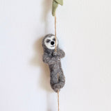 A serene wool felt garland featuring a gray sloth is displayed against a stark white wall. The textured gray wool felt sloth, with a white face, small black nose, and mouth, is shown clinging to a thin, twisted brown rope. Its arms and legs are wrapped around the rope, which extends from a leafy green felt branch at the top.
