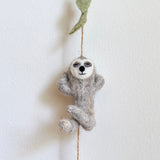 A gray wool felt sloth, part of the Slothful Serenity felt garland, hangs suspended by a natural twine against a white wall. The soft wool felt sloth features a peaceful expression with closed eyes and a gentle smile, its arms raised in a relaxed pose. A small green felt leaf is attached to the twine above the sloth.