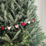 A vibrant Felt Love wool felt garland gracefully hangs on a lush green Christmas tree branch. This charming garland showcases alternating red, blush pink, and white and pink felt balls along with delicate felt hearts, creating a cozy and festive atmosphere.